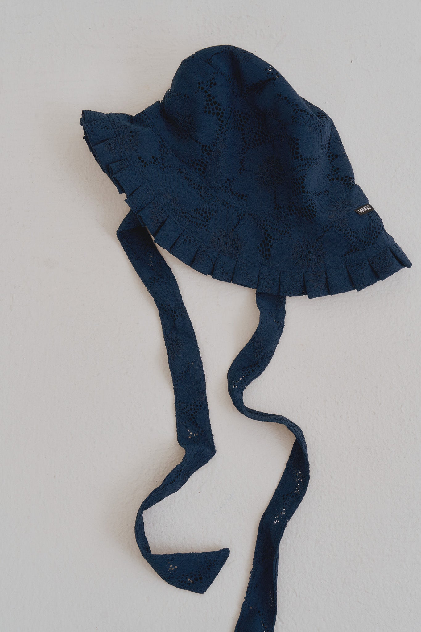 閃靛藍手作蕾絲百褶帽平拍圖，展現細緻罌粟花蕾絲面料、百褶邊緣與可編織的長綁帶設計。
