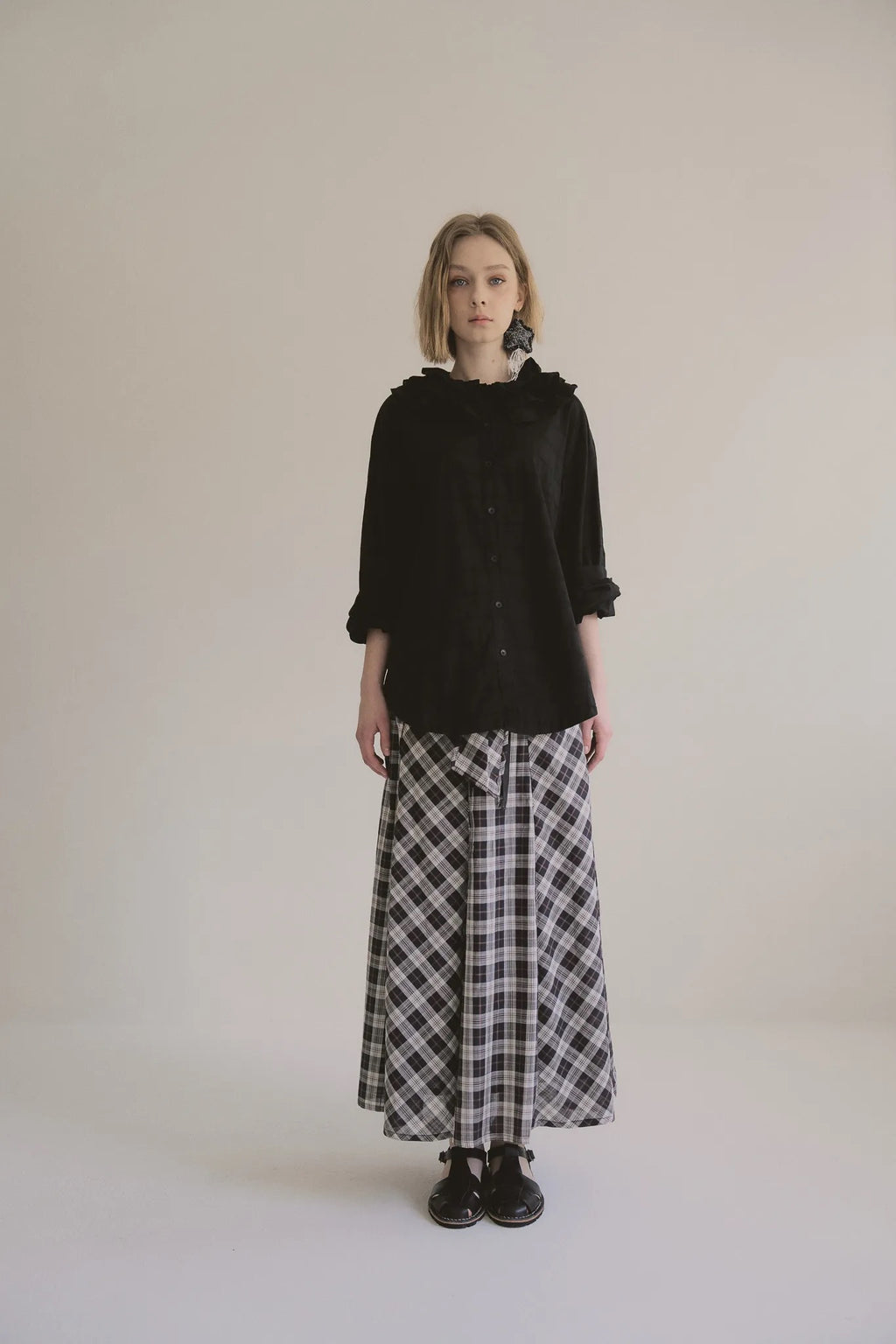 模特兒穿著黑色格紋斜裁綁帶長裙，腰間綁帶與腰釦修飾比例，兩側口袋與裙擺歐干紗營造修長優雅造型。