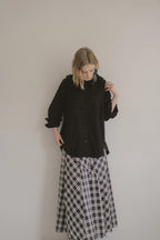 模特兒穿著黑色格紋斜裁綁帶長裙，腰間綁帶與腰釦修飾比例，兩側口袋與裙擺歐干紗營造修長優雅造型。