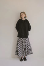 模特兒穿著黑色格紋斜裁綁帶長裙，腰間綁帶與腰釦修飾比例，兩側口袋與裙擺歐干紗營造修長優雅造型。