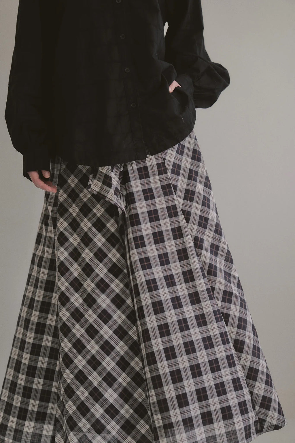 模特兒穿著黑色格紋斜裁綁帶長裙，腰間綁帶與腰釦修飾比例，兩側口袋與裙擺歐干紗營造修長優雅造型。