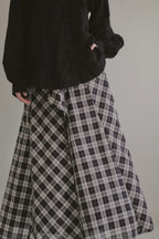 模特兒穿著黑色格紋斜裁綁帶長裙，腰間綁帶與腰釦修飾比例，兩側口袋與裙擺歐干紗營造修長優雅造型。