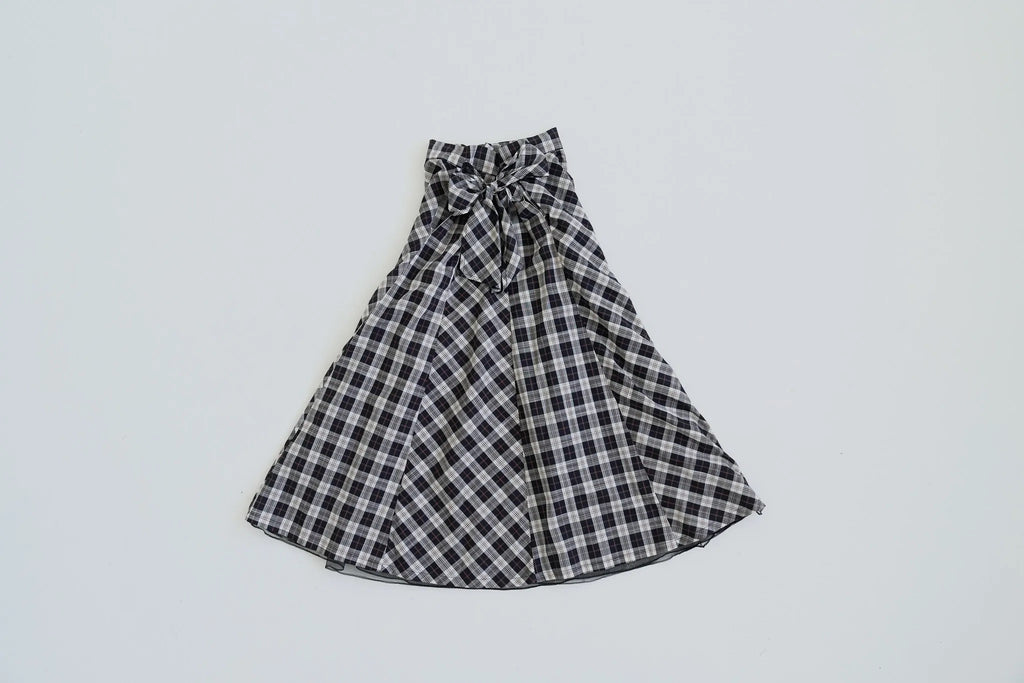 模特兒穿著黑色格紋斜裁綁帶長裙，腰間綁帶與腰釦修飾比例，兩側口袋與裙擺歐干紗營造修長優雅造型。