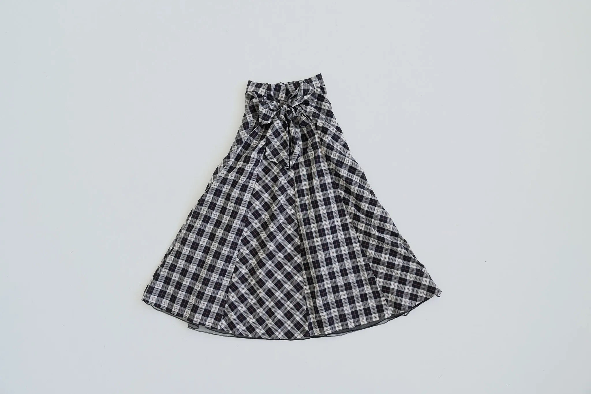 模特兒穿著黑色格紋斜裁綁帶長裙，腰間綁帶與腰釦修飾比例，兩側口袋與裙擺歐干紗營造修長優雅造型。
