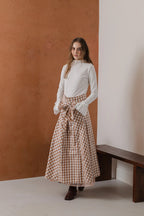 模特兒穿著紅棕色格紋斜裁綁帶長裙，腰間綁帶與腰釦修飾比例，兩側口袋與裙擺歐干紗營造修長優雅造型。