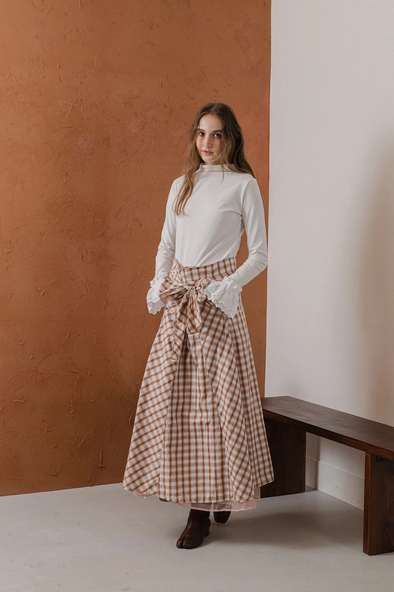 模特兒穿著紅棕色格紋斜裁綁帶長裙，腰間綁帶與腰釦修飾比例，兩側口袋與裙擺歐干紗營造修長優雅造型。