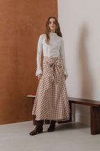 模特兒穿著紅棕色格紋斜裁綁帶長裙，腰間綁帶與腰釦修飾比例，兩側口袋與裙擺歐干紗營造修長優雅造型。