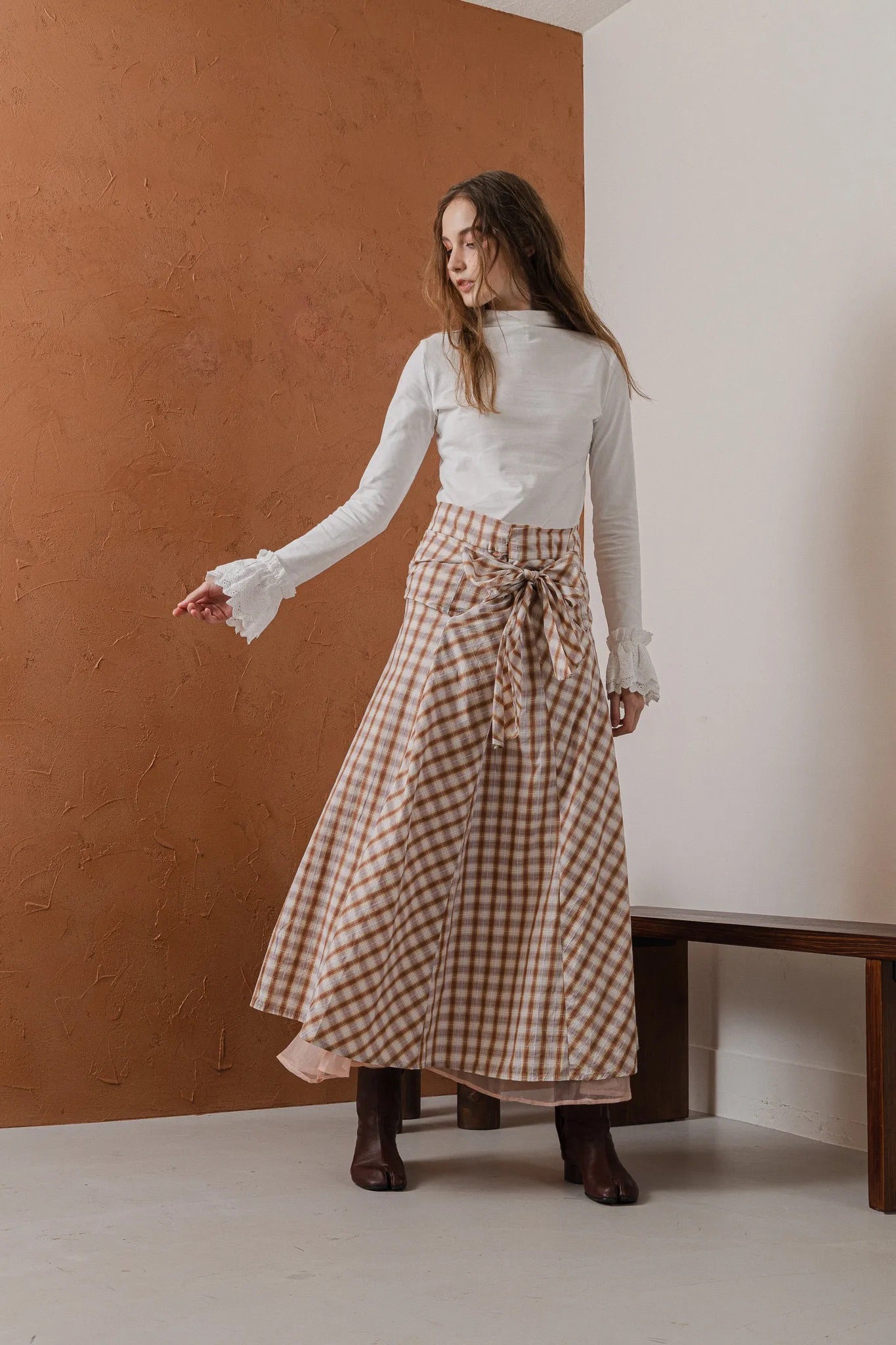 模特兒穿著紅棕色格紋斜裁綁帶長裙，腰間綁帶與腰釦修飾比例，兩側口袋與裙擺歐干紗營造修長優雅造型。