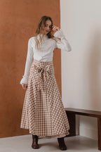 模特兒穿著紅棕色格紋斜裁綁帶長裙，腰間綁帶與腰釦修飾比例，兩側口袋與裙擺歐干紗營造修長優雅造型。