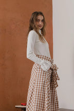 模特兒穿著紅棕色格紋斜裁綁帶長裙，腰間綁帶與腰釦修飾比例，兩側口袋與裙擺歐干紗營造修長優雅造型。
