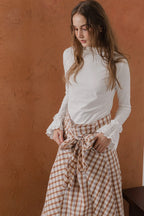 模特兒穿著紅棕色格紋斜裁綁帶長裙，腰間綁帶與腰釦修飾比例，兩側口袋與裙擺歐干紗營造修長優雅造型。