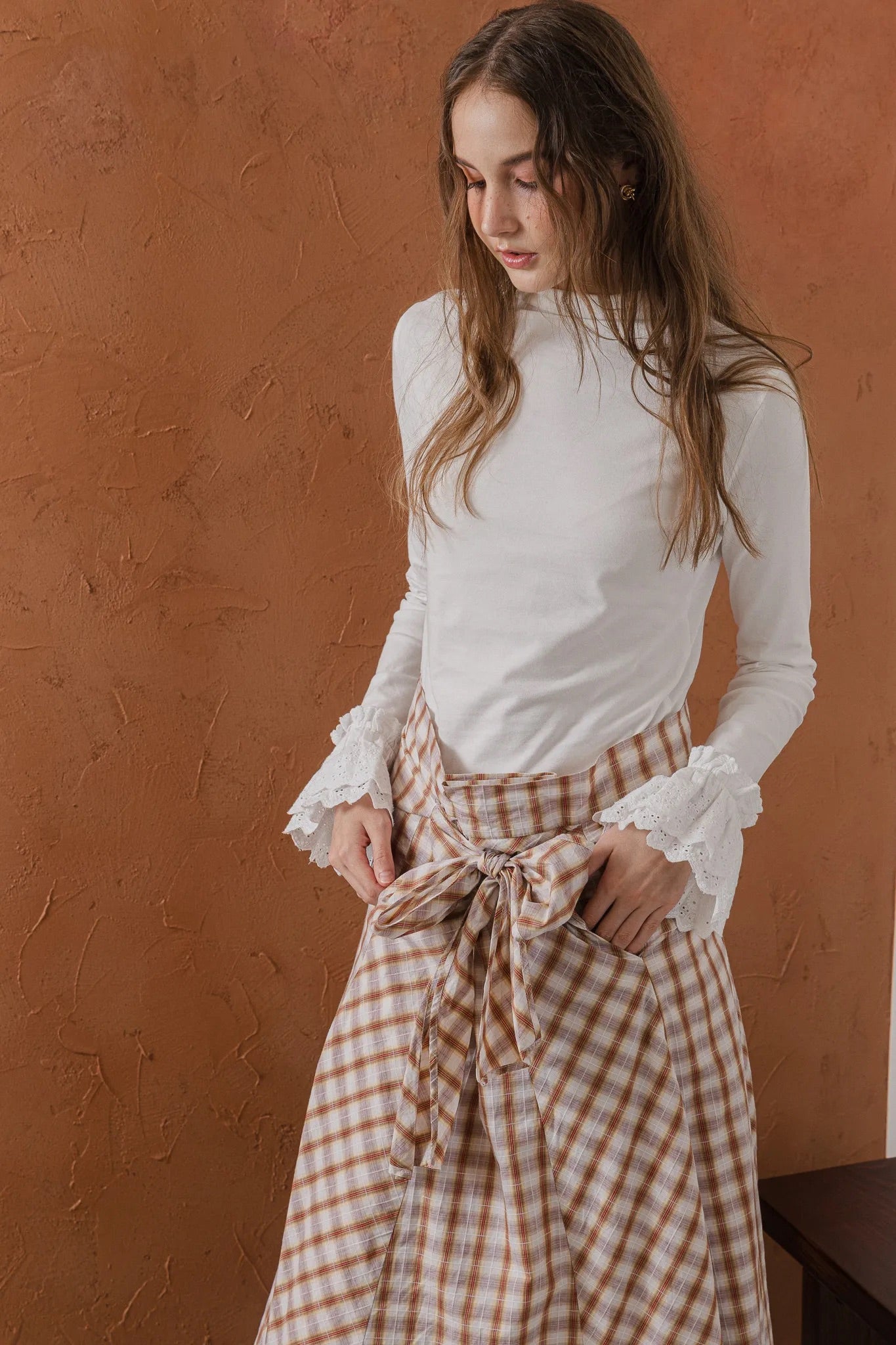 模特兒穿著紅棕色格紋斜裁綁帶長裙，腰間綁帶與腰釦修飾比例，兩側口袋與裙擺歐干紗營造修長優雅造型。