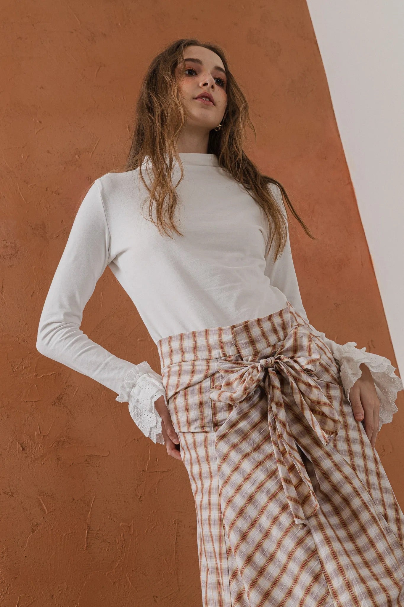 模特兒穿著紅棕色格紋斜裁綁帶長裙，腰間綁帶與腰釦修飾比例，兩側口袋與裙擺歐干紗營造修長優雅造型。
