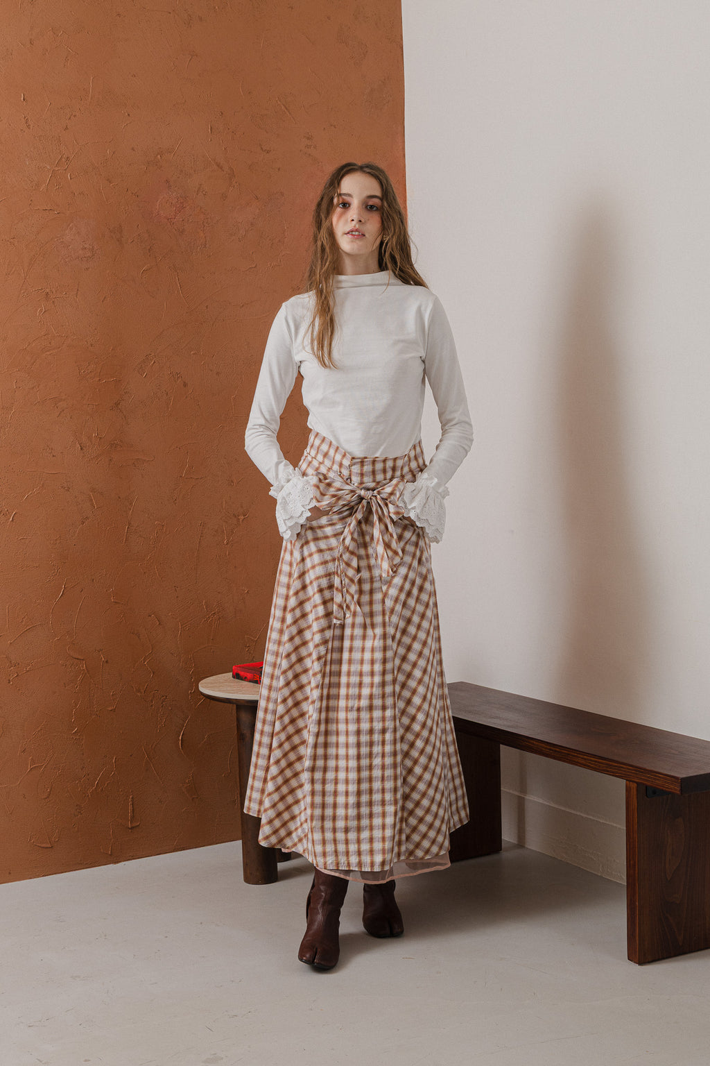模特兒穿著紅棕色格紋斜裁綁帶長裙，腰間綁帶與腰釦修飾比例，兩側口袋與裙擺歐干紗營造修長優雅造型。