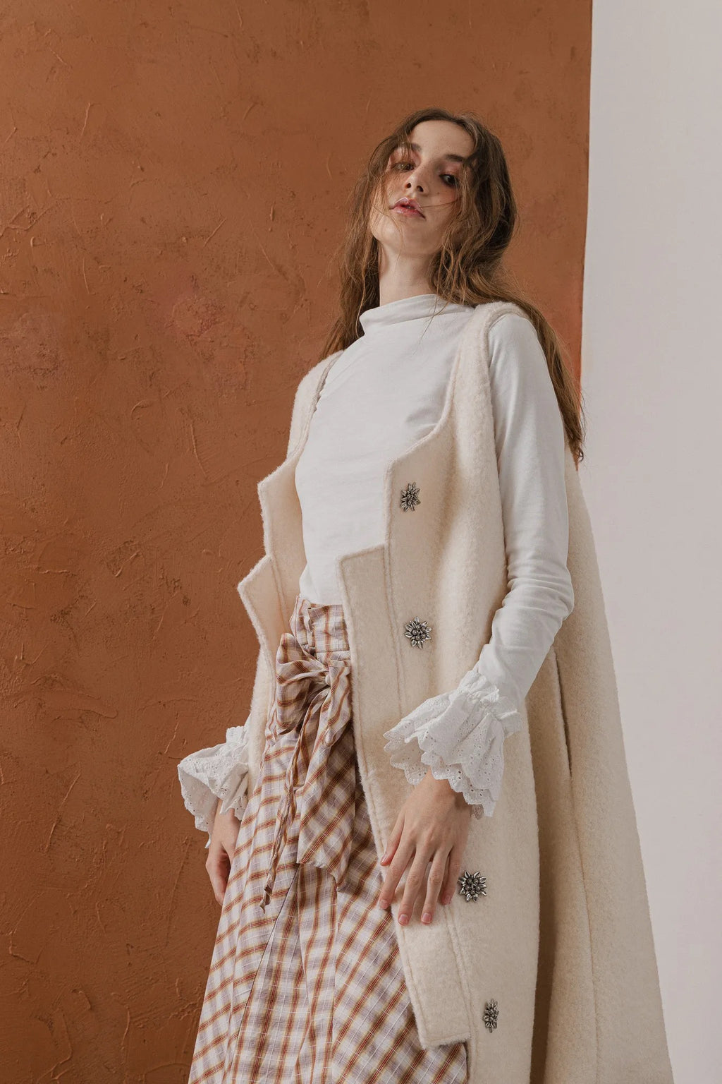 模特兒穿著燕麥白羊絨背心洋裝外套，柔軟羊絨面料搭配立體花釦、後開衩與側袋設計，展現優雅多變造型。