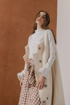 模特兒穿著燕麥白羊絨背心洋裝外套，柔軟羊絨面料搭配立體花釦、後開衩與側袋設計，展現優雅多變造型。