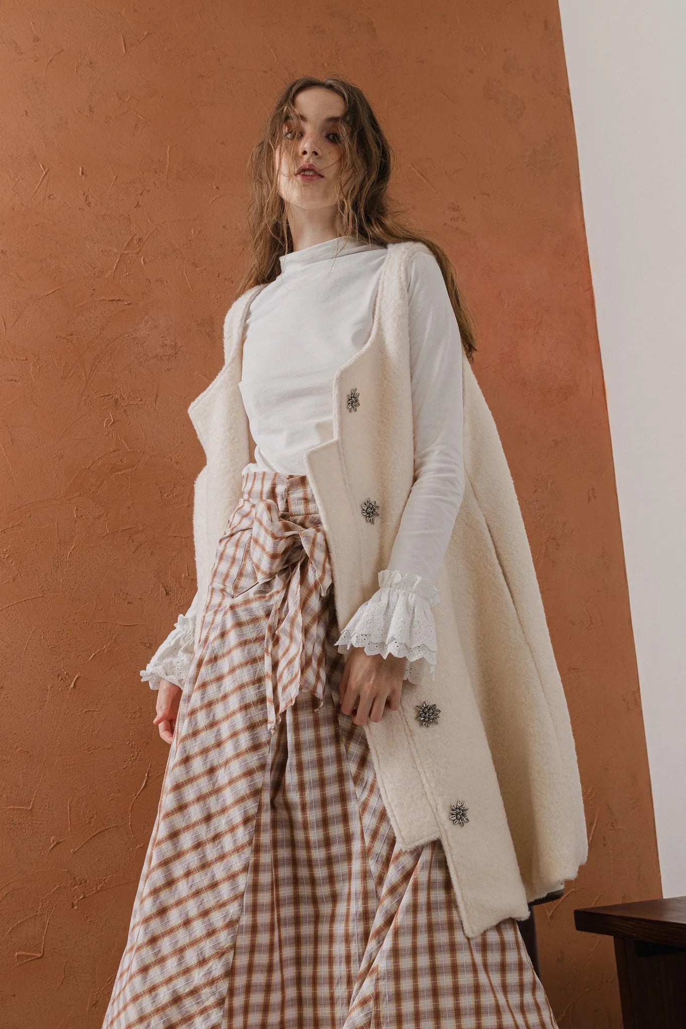 模特兒穿著燕麥白羊絨背心洋裝外套，柔軟羊絨面料搭配立體花釦、後開衩與側袋設計，展現優雅多變造型。