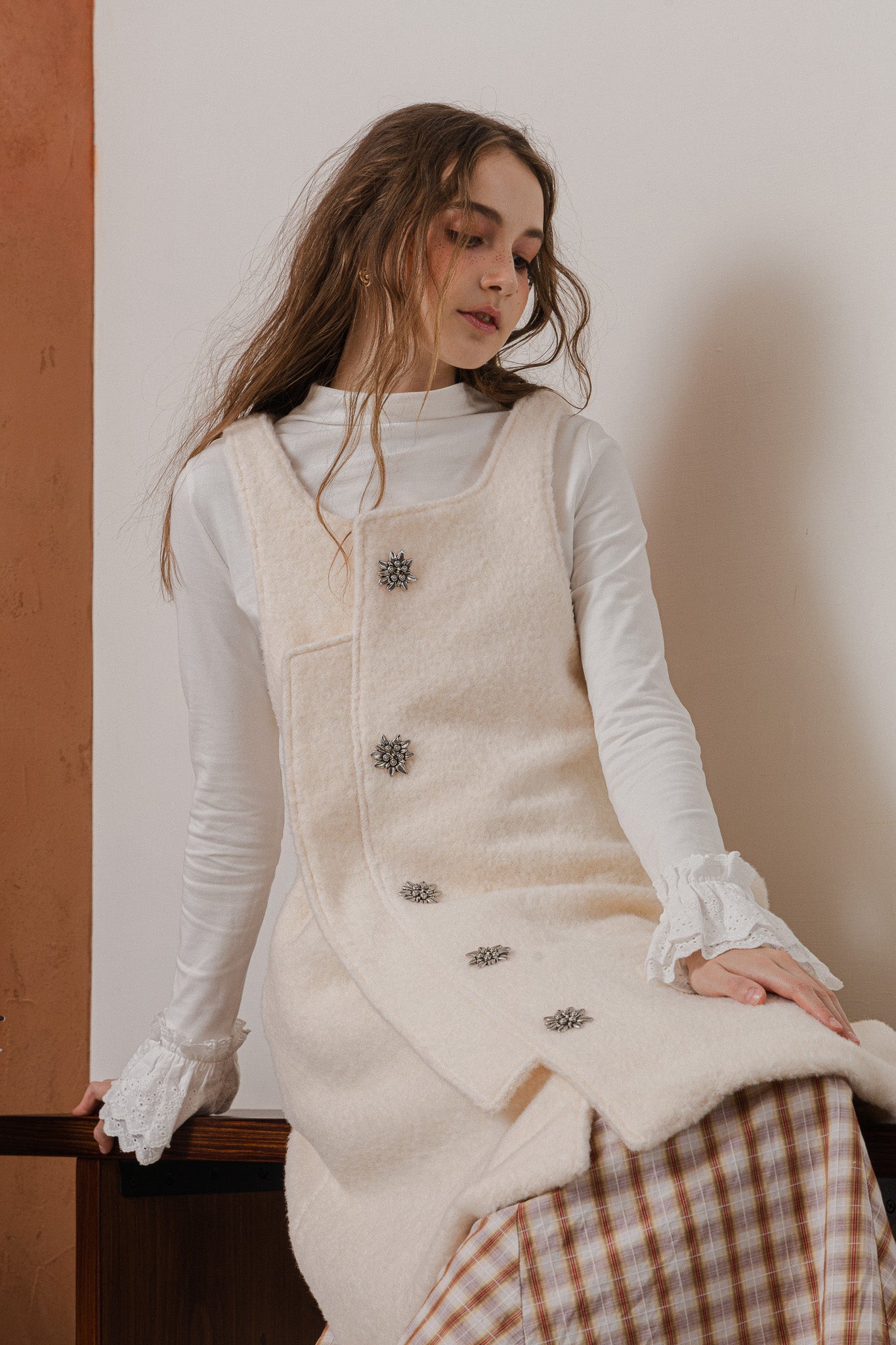 模特兒穿著燕麥白羊絨背心洋裝外套，柔軟羊絨面料搭配立體花釦、後開衩與側袋設計，展現優雅多變造型。