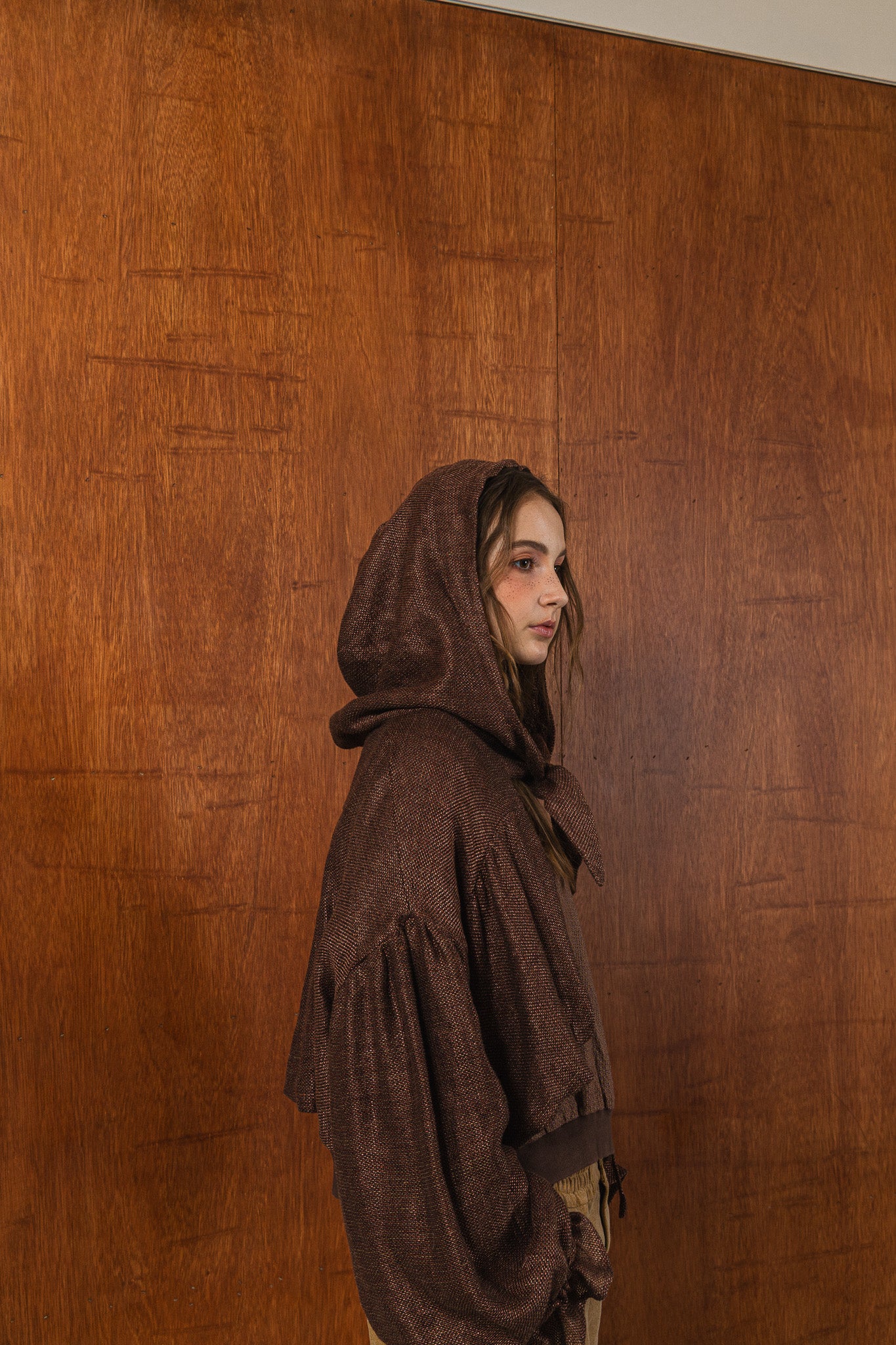 模特兒穿著光澤棕色童話感連帽外套，光澤針織面料與前綁帶設計搭配前短後長剪裁，呈現優雅又帶點叛逆氛圍的造型。