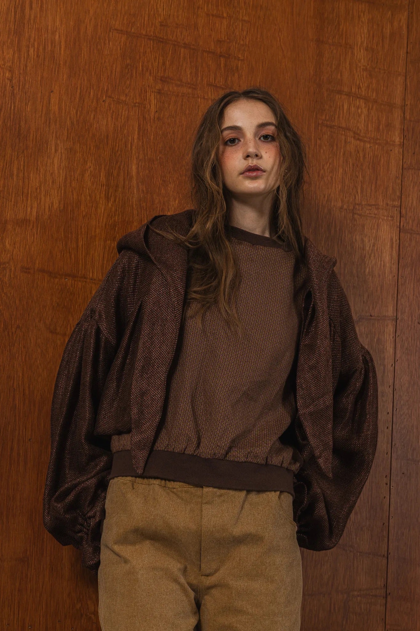 模特兒穿著光澤棕色童話感連帽外套，光澤針織面料與前綁帶設計搭配前短後長剪裁，呈現優雅又帶點叛逆氛圍的造型。