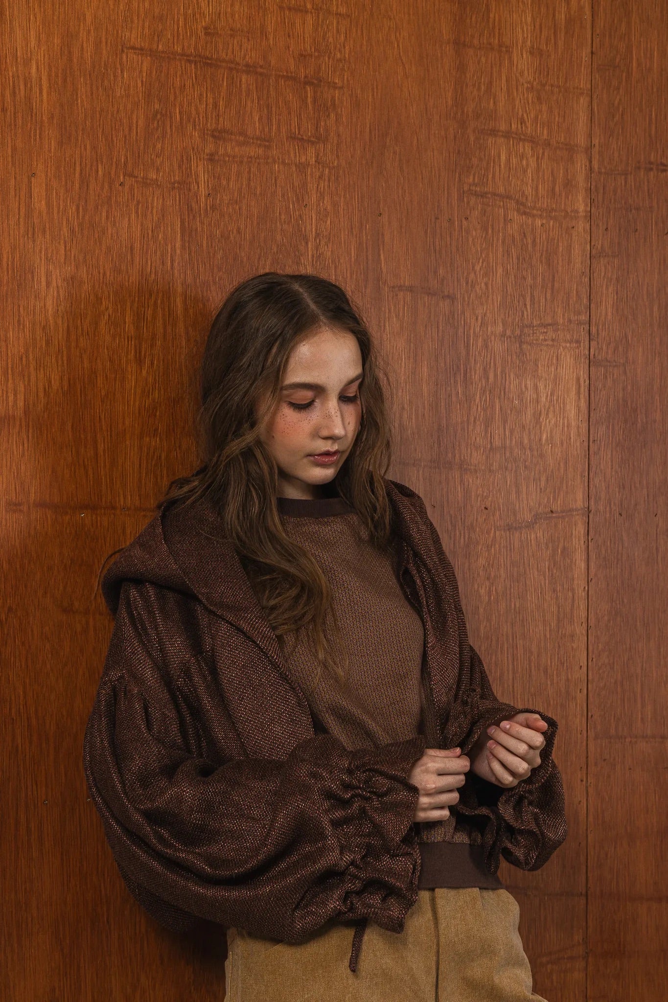 模特兒穿著光澤棕色童話感連帽外套，光澤針織面料與前綁帶設計搭配前短後長剪裁，呈現優雅又帶點叛逆氛圍的造型。