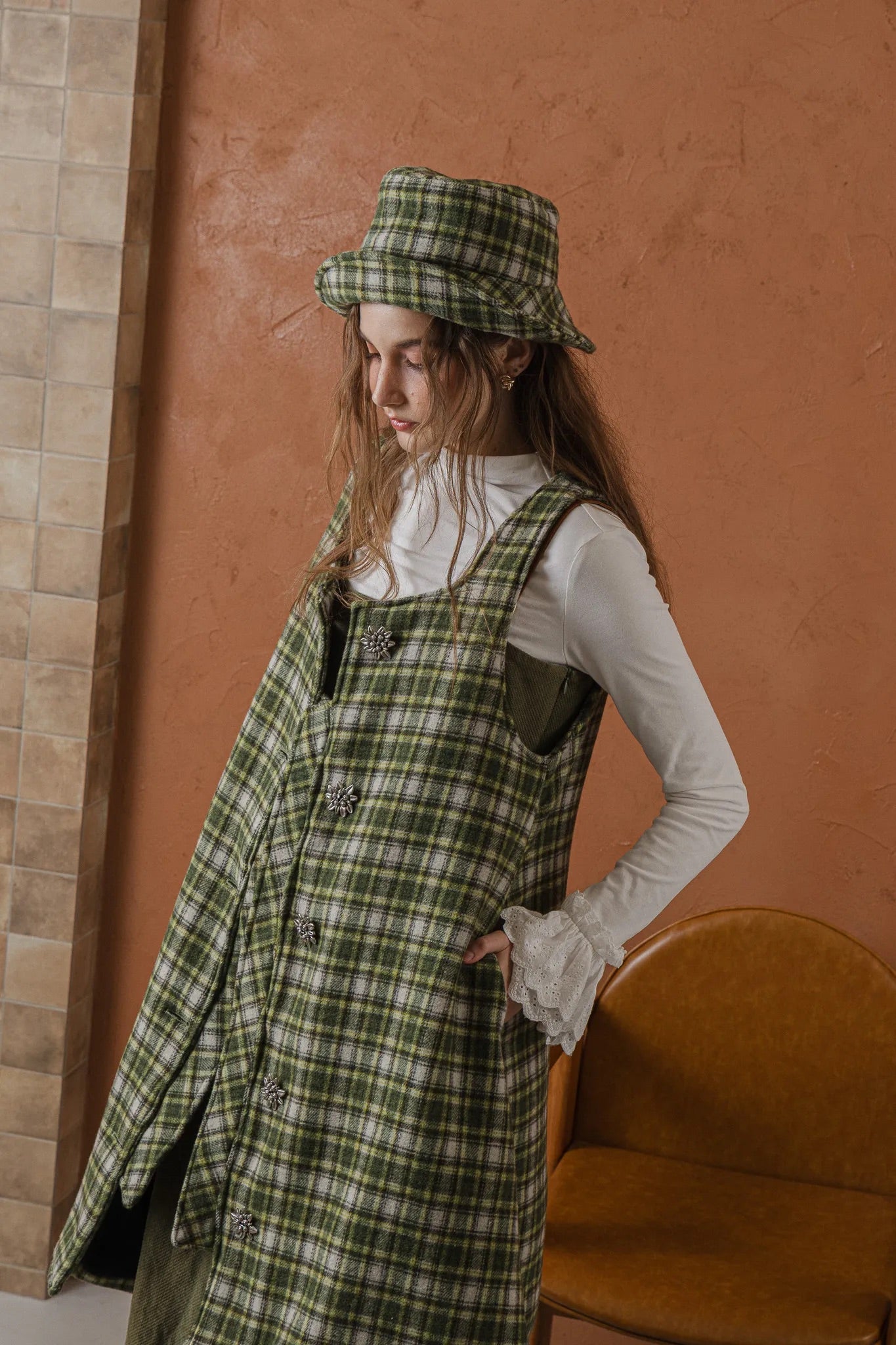 模特兒穿著羊毛格紋背心洋裝外套，經典學院風格紋搭配獨特門襟與手縫立體花釦，後開衩與側口袋設計呈現優雅又隨性的造型。